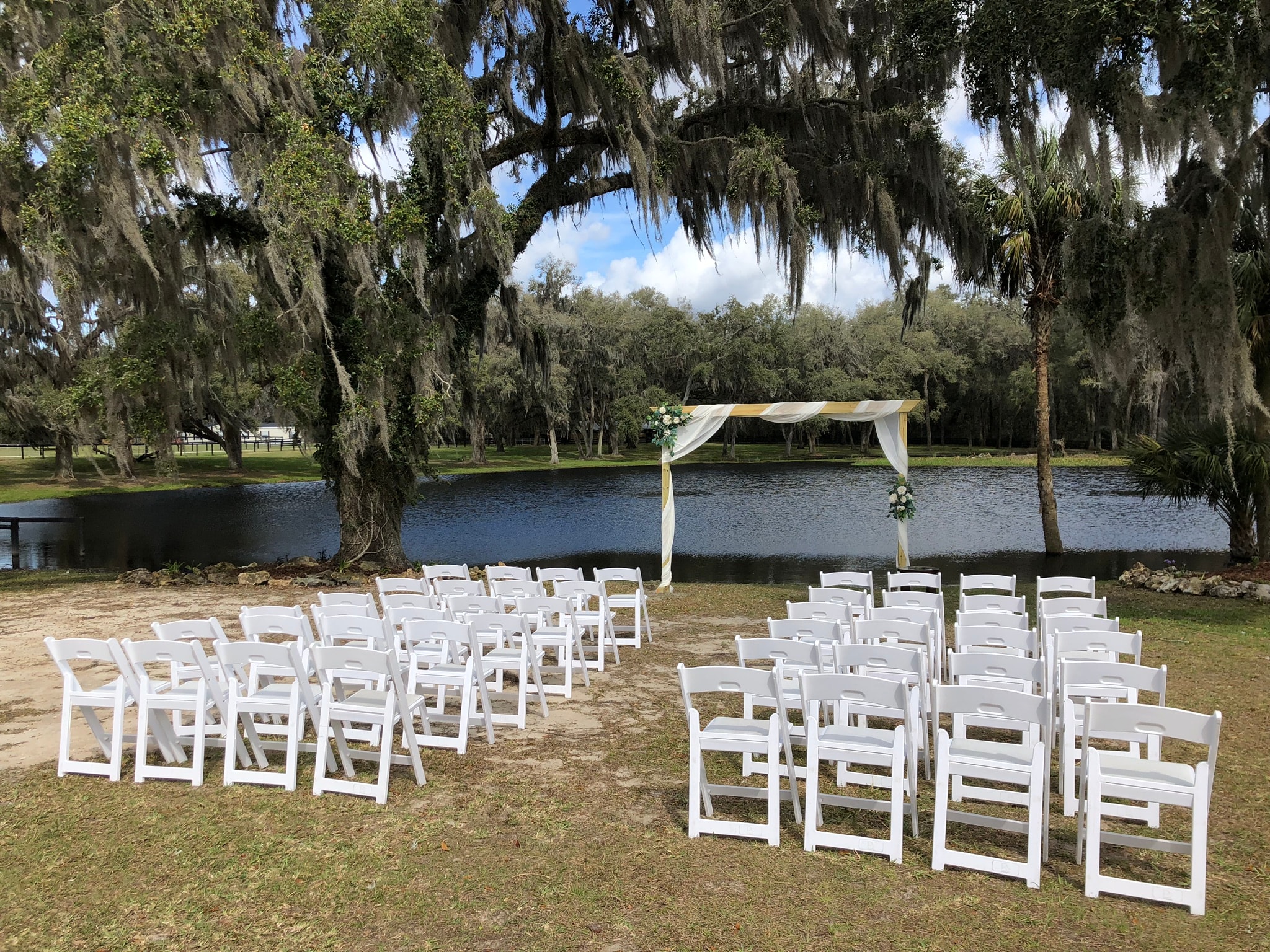 Outdoor ceremony setup with white chairs and draped arch overlooking the ranch lake under Spanish moss oaks