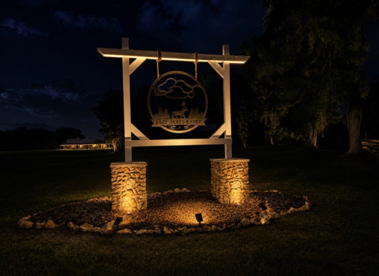 Lazy Acres Ranch illuminated entrance sign glowing at night with stone pillars and landscape lighting