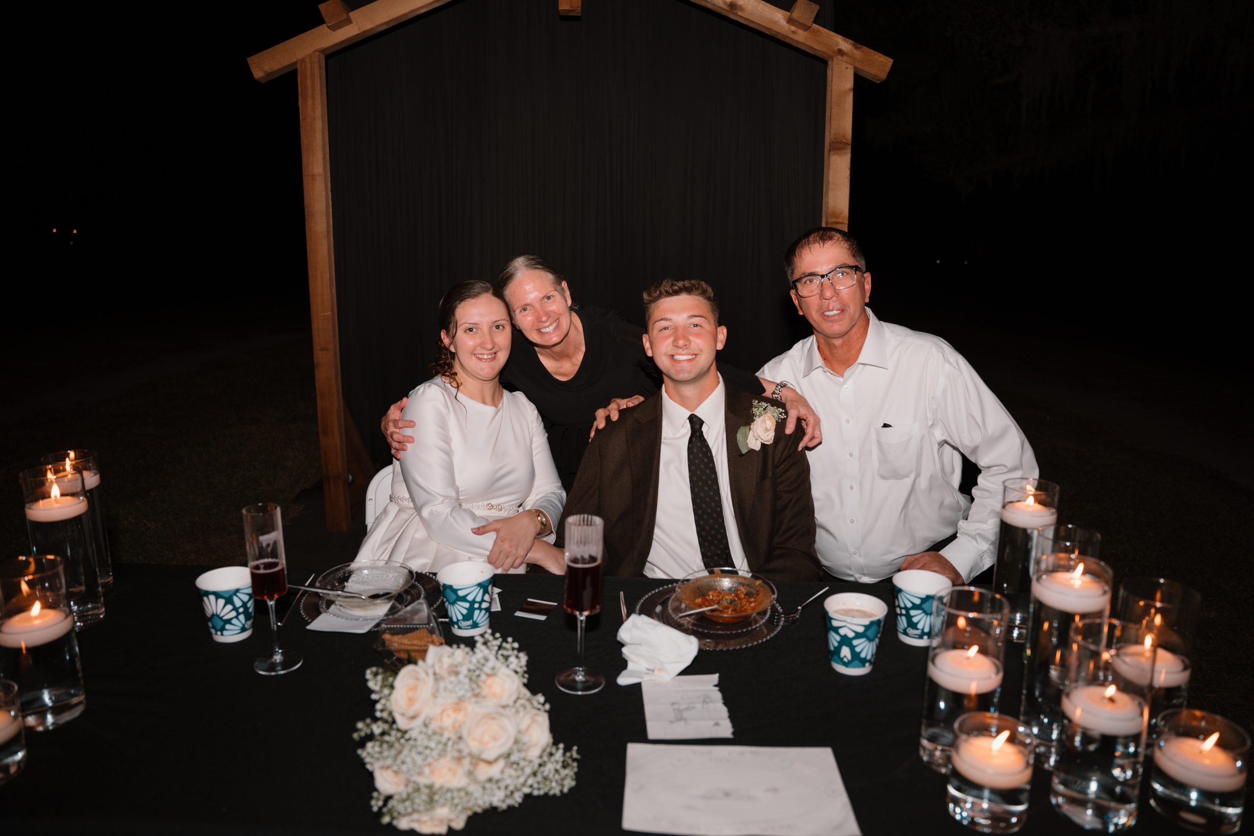 Four guests smiling at a candlelit reception table with white roses, floating candles, and the wooden arbor behind them