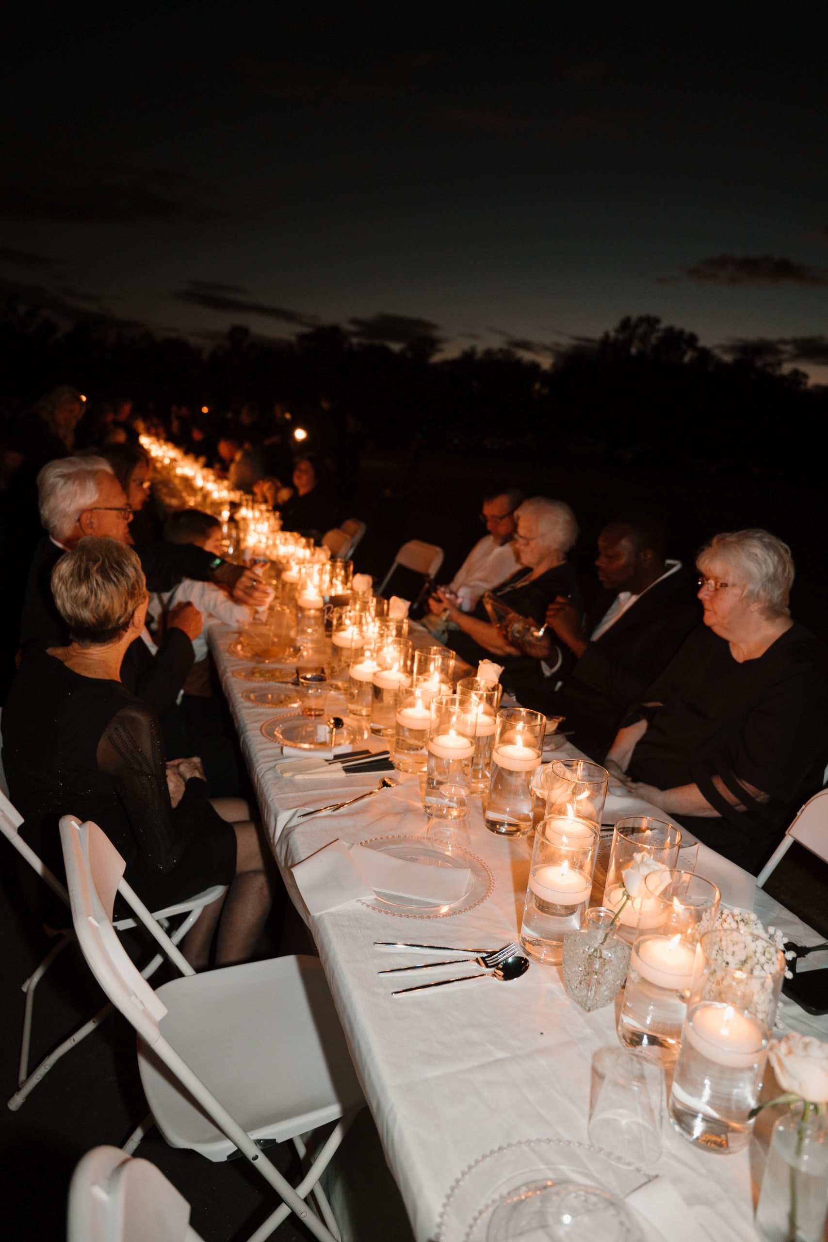 Guests in formal attire dining at a long candlelit outdoor table stretching into the dusk horizon