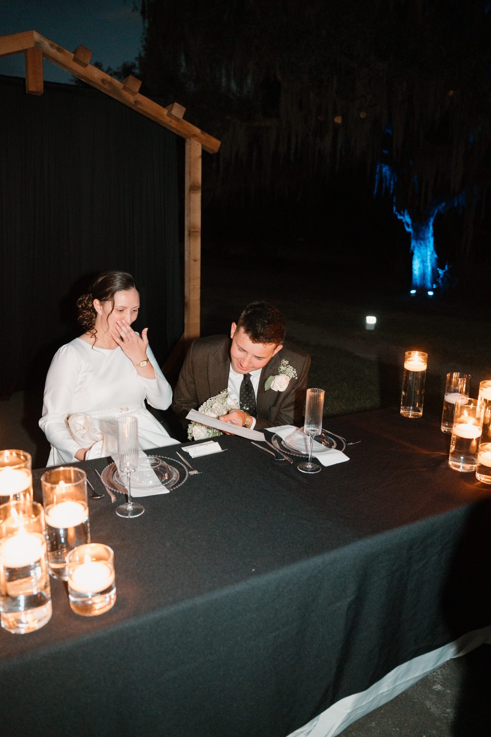 Couple at a candlelit sweetheart table at night under the wooden arbor with blue-lit oak trees in the background