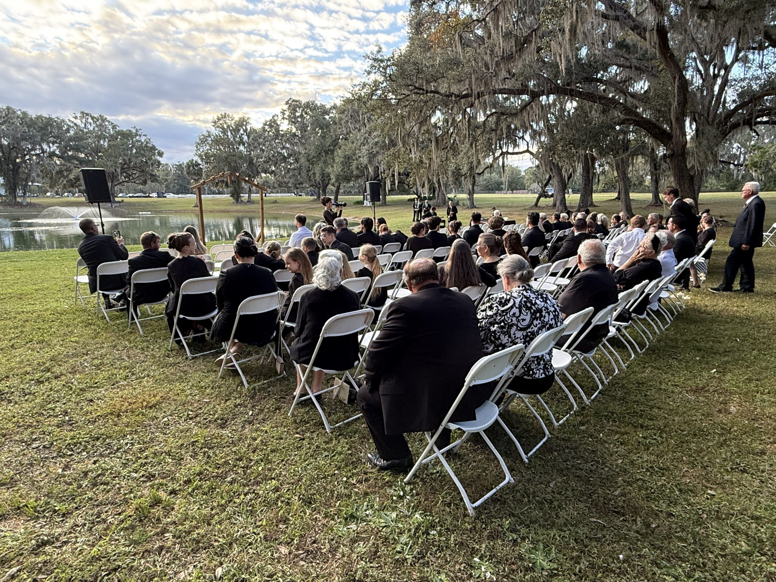 Guests seated in white chairs for an outdoor ceremony by the lake under Spanish moss oak trees