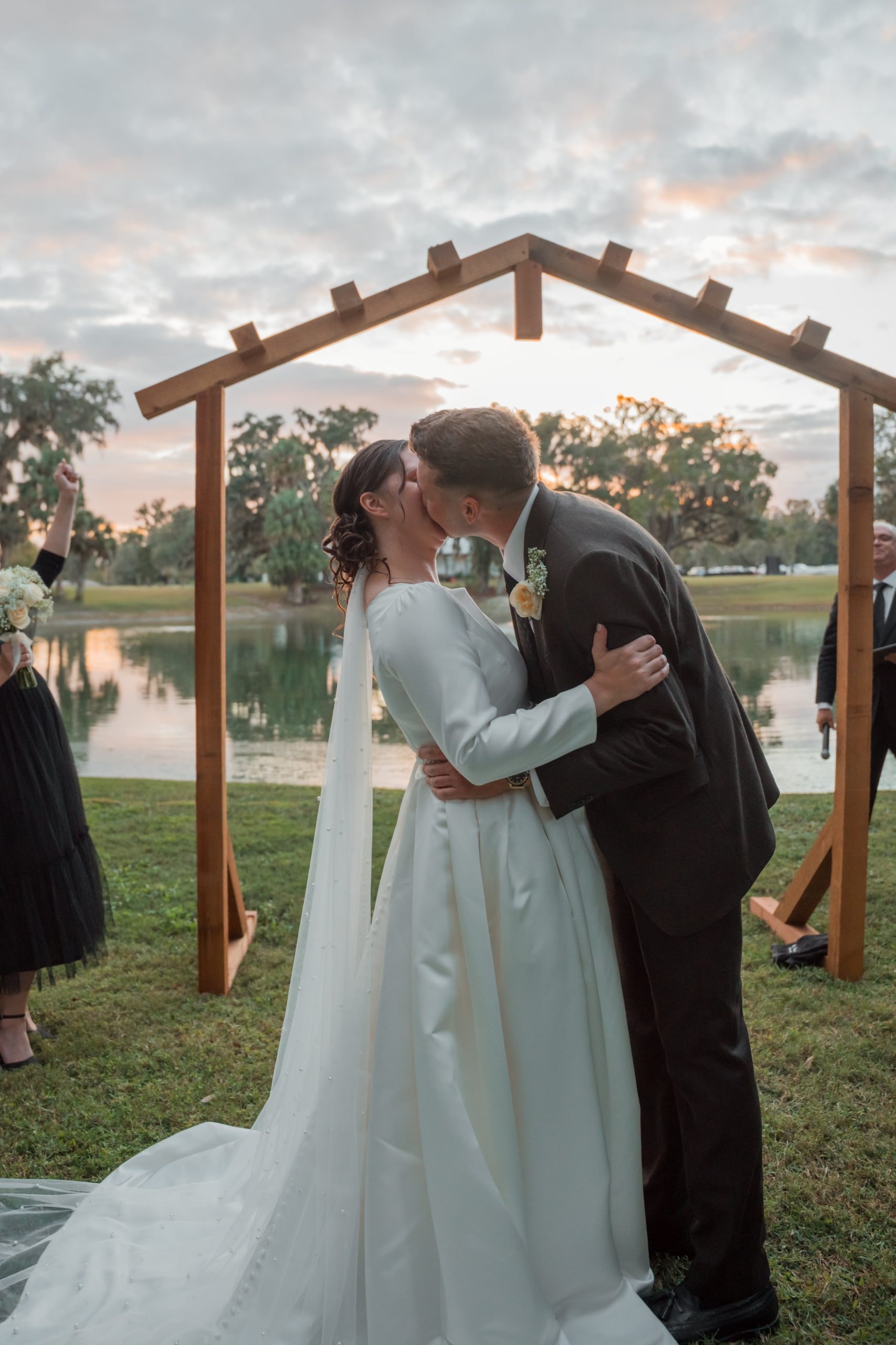 Couple sharing their first kiss under the wooden arbor with the lake and a pastel sunset sky glowing behind them