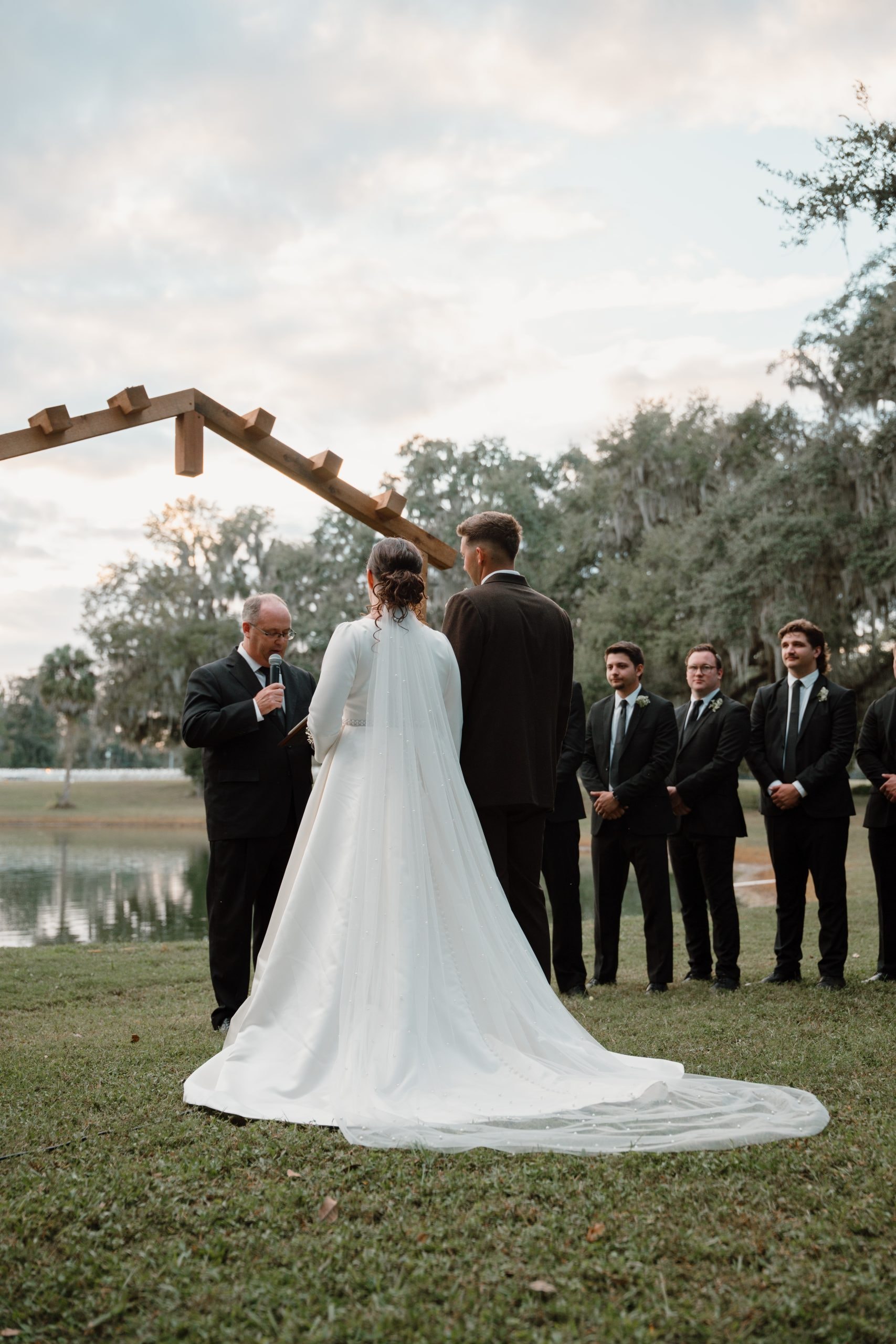 Bride and groom exchanging vows under the wooden arbor at dusk with the lake and Spanish moss trees behind them