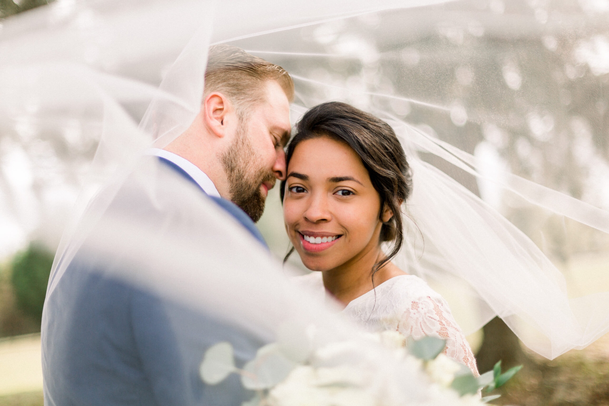 Romantic close-up of bride smiling under flowing veil with groom on the ranch grounds