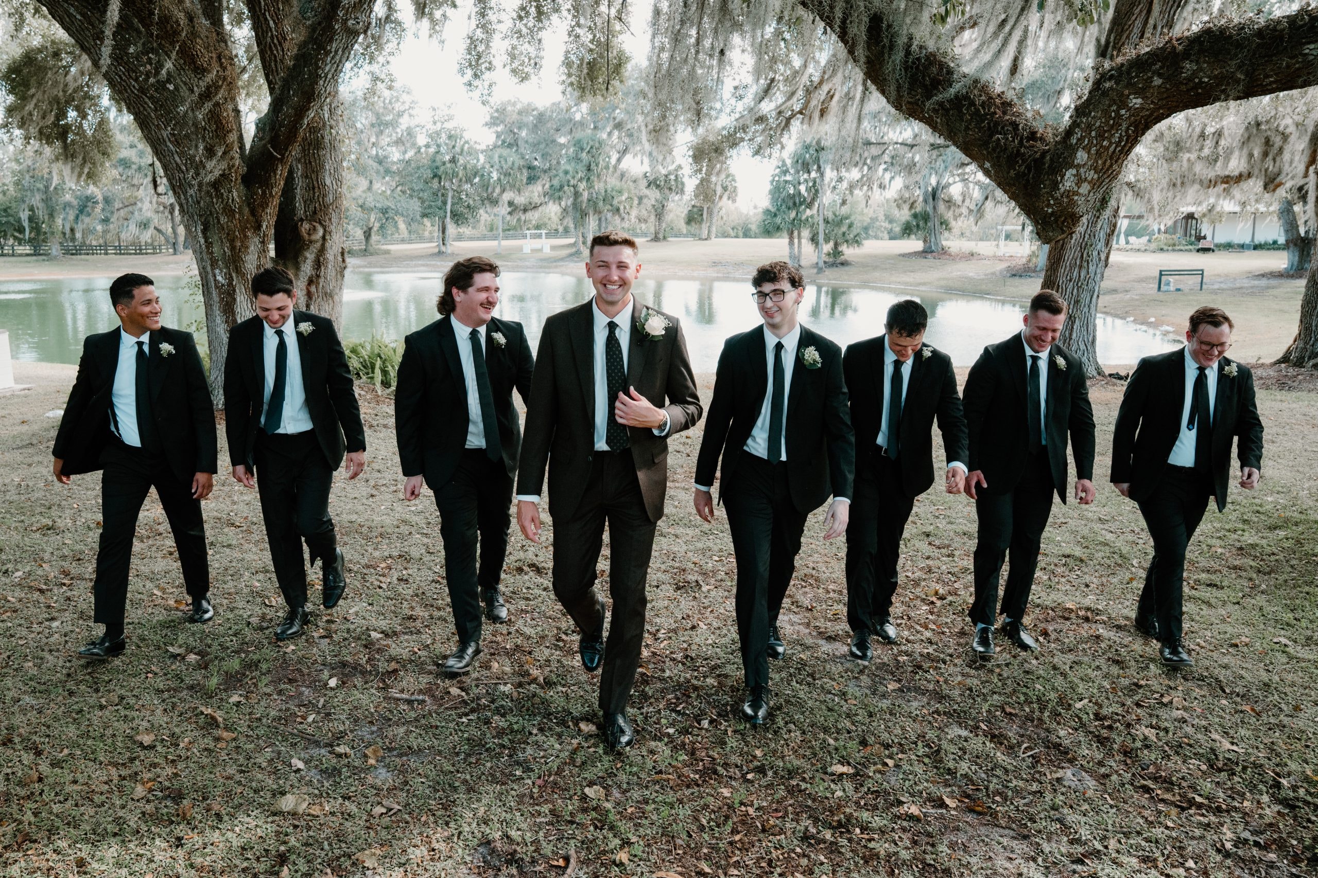 Groomsmen walking together along the lakeside grounds under oak trees