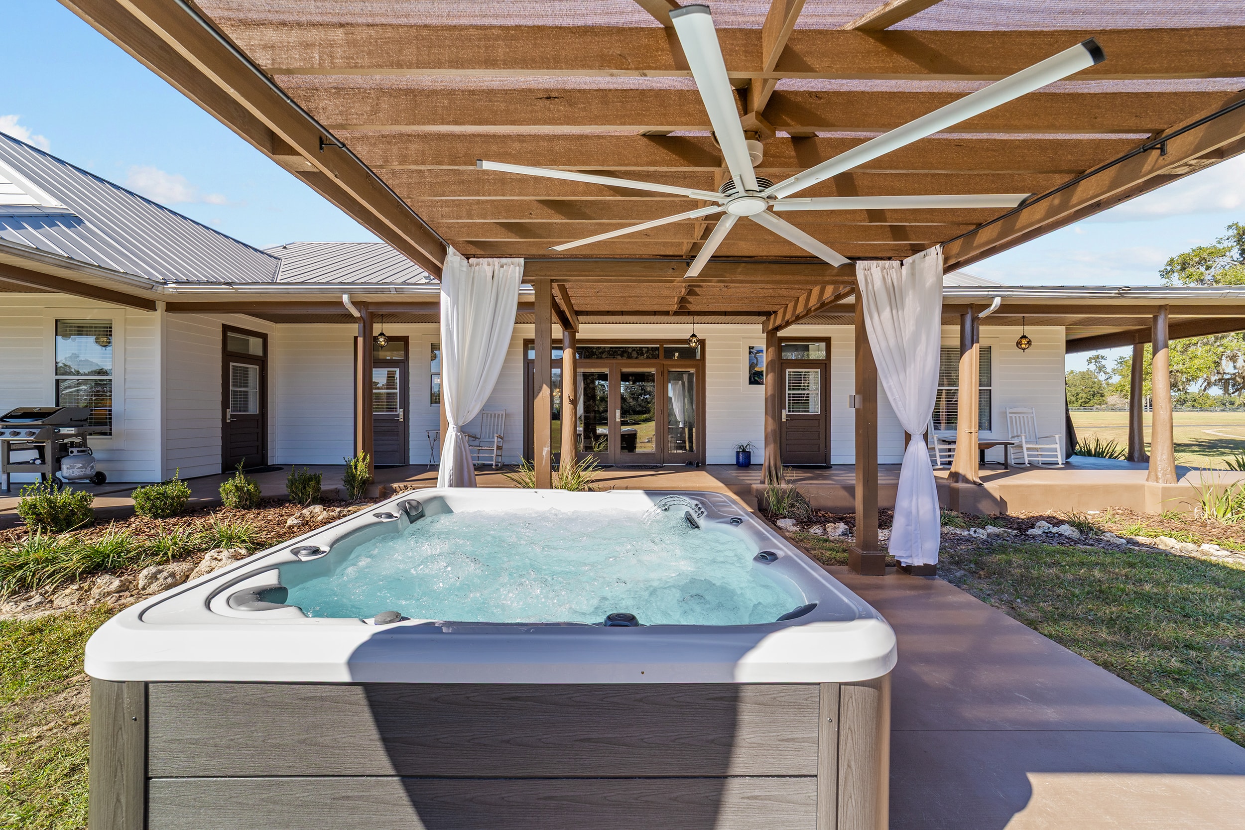 Bubbling hot tub under a shaded pergola with large ceiling fan and white drape curtains on the estate back patio