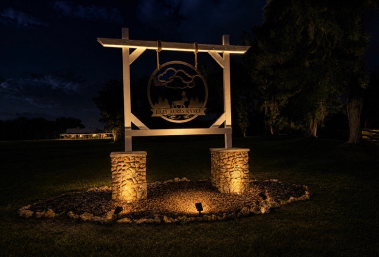Lazy Acres Ranch illuminated entrance sign glowing at night with stone pillars and landscape lighting